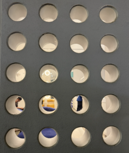 A textured gray wall featuring a rigid grid of deep circular cutouts that reveal fragmented views of a brightly lit interior space with people and colorful furniture.
