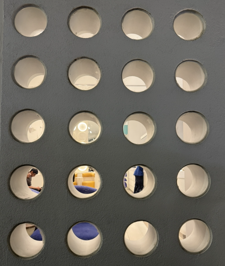 A textured gray wall featuring a rigid grid of deep circular cutouts that reveal fragmented views of a brightly lit interior space with people and colorful furniture.