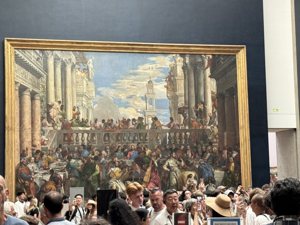 Paolo Veronese's colossal The Wedding Feast at Cana hanging directly opposite the Mona Lisa, being photographed by a large dense crowd of museum tourists in the foreground.