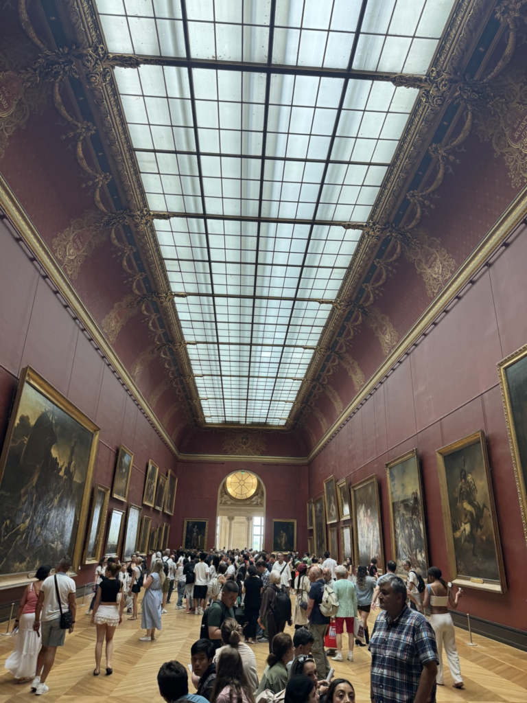 A wide perspective down a grand crowded museum gallery featuring deep red walls, classic parquet flooring, a massive glass skylight, and monumental 19th-century French paintings in large gold frames.