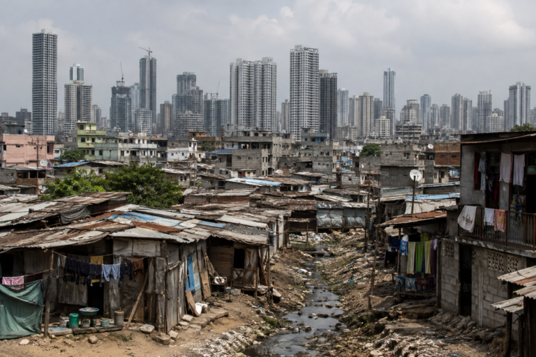 A polluted stream runs through a densely packed slum with makeshift metal and brick houses in the foreground, contrasting sharply with a modern skyline of tall skyscrapers in the background.