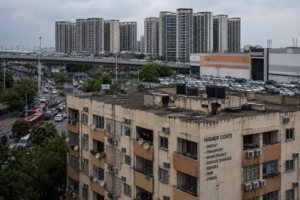 An older, weathered apartment building with the words "HIGHER COSTS" and a list of everyday expenses painted on its side, standing in front of a busy highway, a crowded parking lot, and towering modern high-rises under an overcast sky.