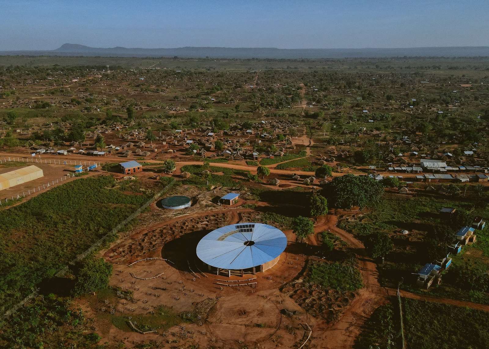 Aerial view of the circular Bidi Bidi Performing Arts Centre, showing its distinctive funnel-shaped roof amid a vast African settlement in Uganda.