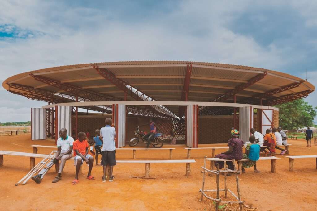 People gather on wooden benches at the Bidi Bidi Performing Arts Centre, with a man on a motorcycle seen through the open earth brick and steel structure.