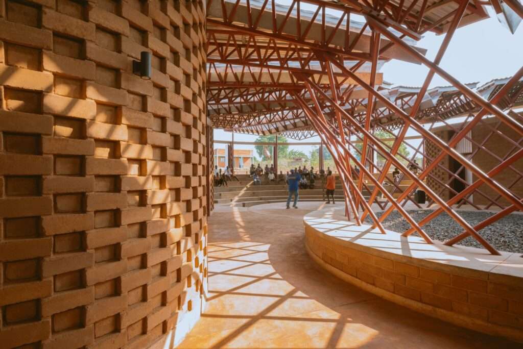 Dramatic geometric shadows from a perforated earth brick wall fall across the floor, looking towards a performance on a circular stage within the Bidi Bidi Performing Arts Centre.