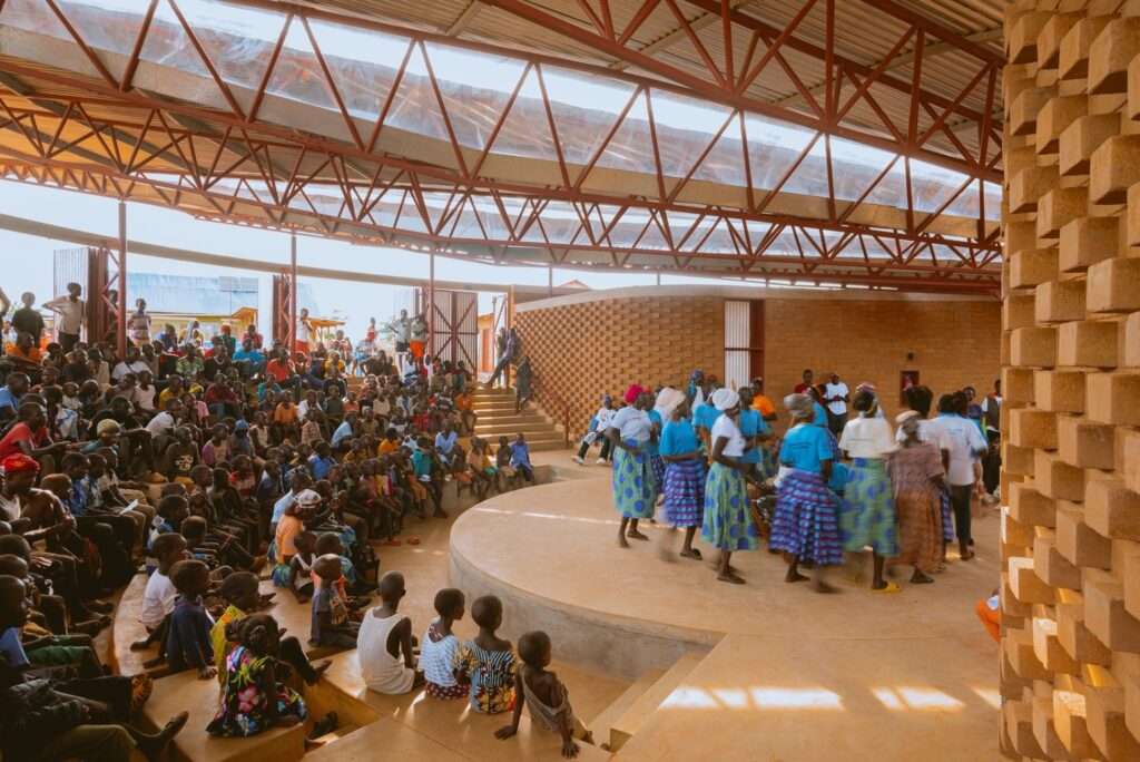 A large audience is seated on earth steps, watching a traditional dance performance in vibrant clothing on a central circular stage at the Bidi Bidi Performing Arts Centre.