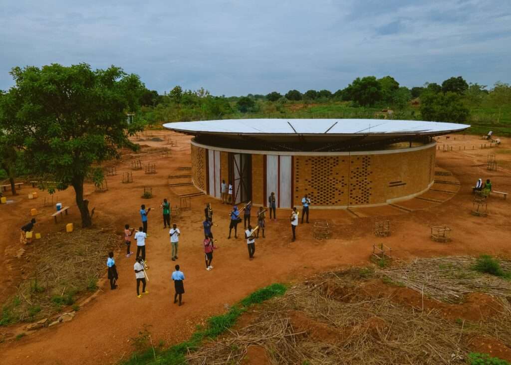 An aerial view of the Bidi Bidi Performing Arts Centre and its water-collecting roof, with a marching band in blue and white uniforms seen in the foreground on the earth paths.