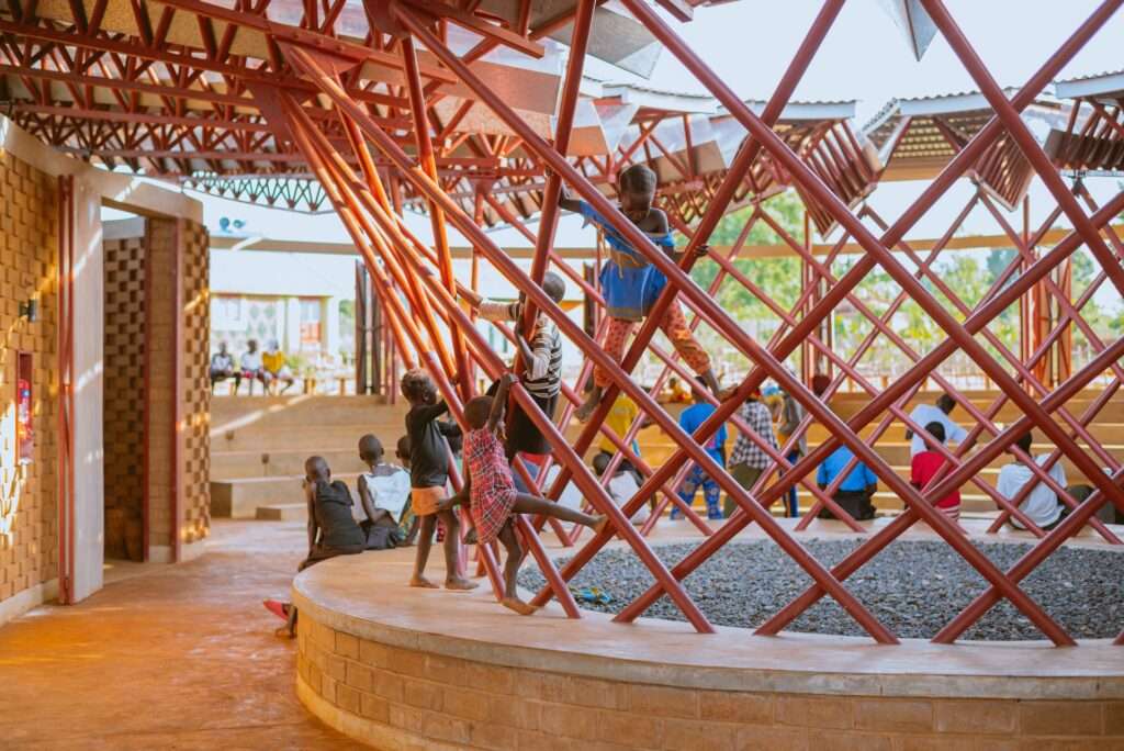 Children climb on a steel pipe structure in a gravel pit inside the Bidi Bidi Performing Arts Centre, with an audience and performance visible beyond.