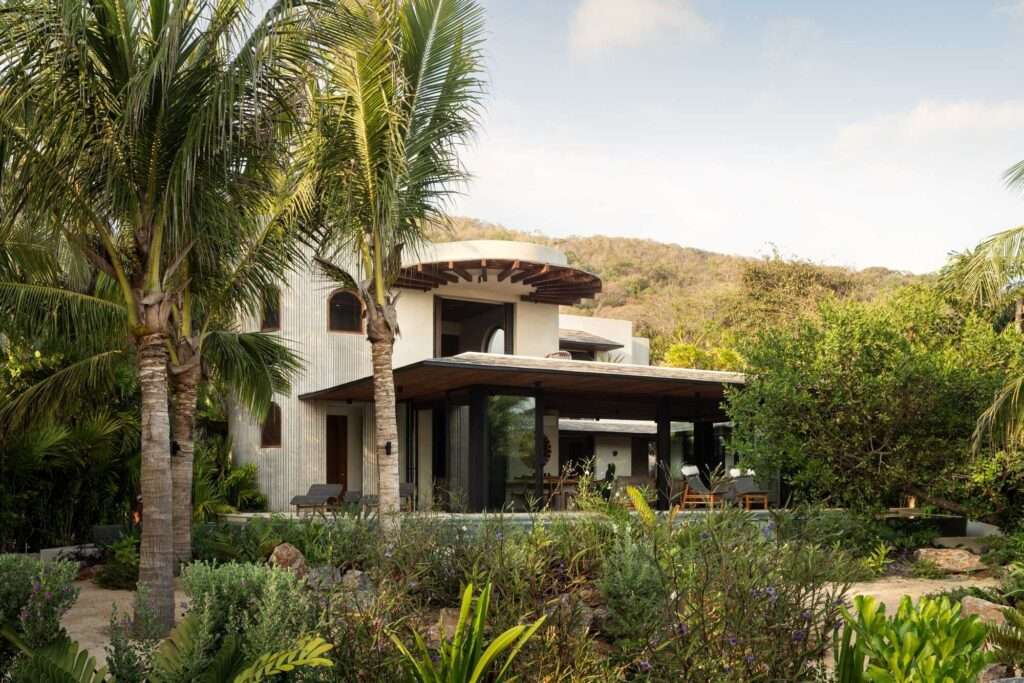 Exterior view of the Pura Vida residential project featuring ribbed concrete walls, arched windows, and a wooden terrace surrounded by lush tropical palm trees.