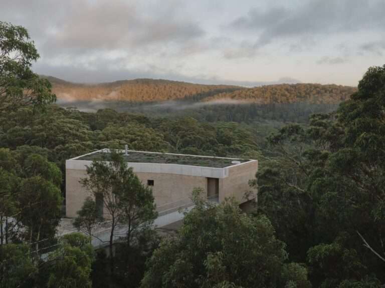 An aerial view of EA House's brick facade and green roof nestled within dense, green bushland, with misty forested hills in the distance at dusk.