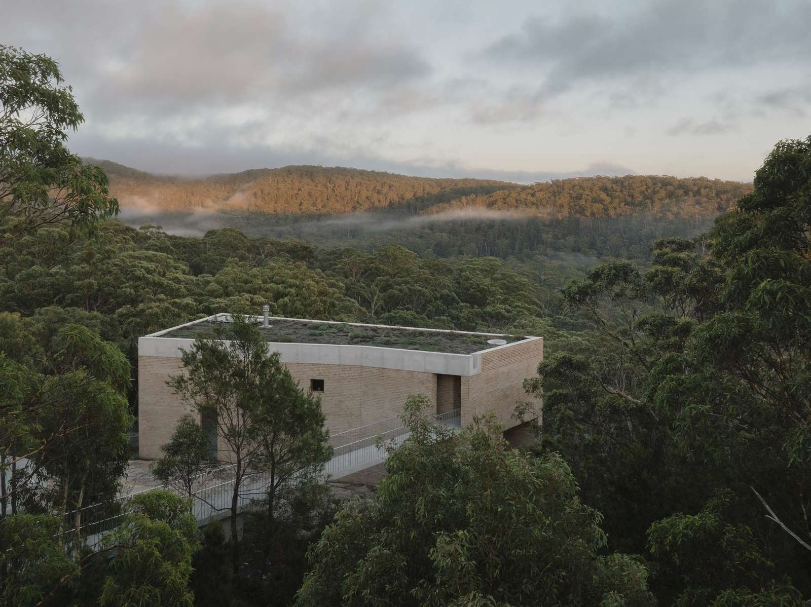 An aerial view of EA House's brick facade and green roof nestled within dense, green bushland, with misty forested hills in the distance at dusk.