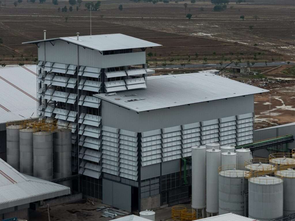 Aerial shot of an industrial rice bran oil refinery facility featuring corrugated metal facade louvers and storage silos set against open agricultural fields.
