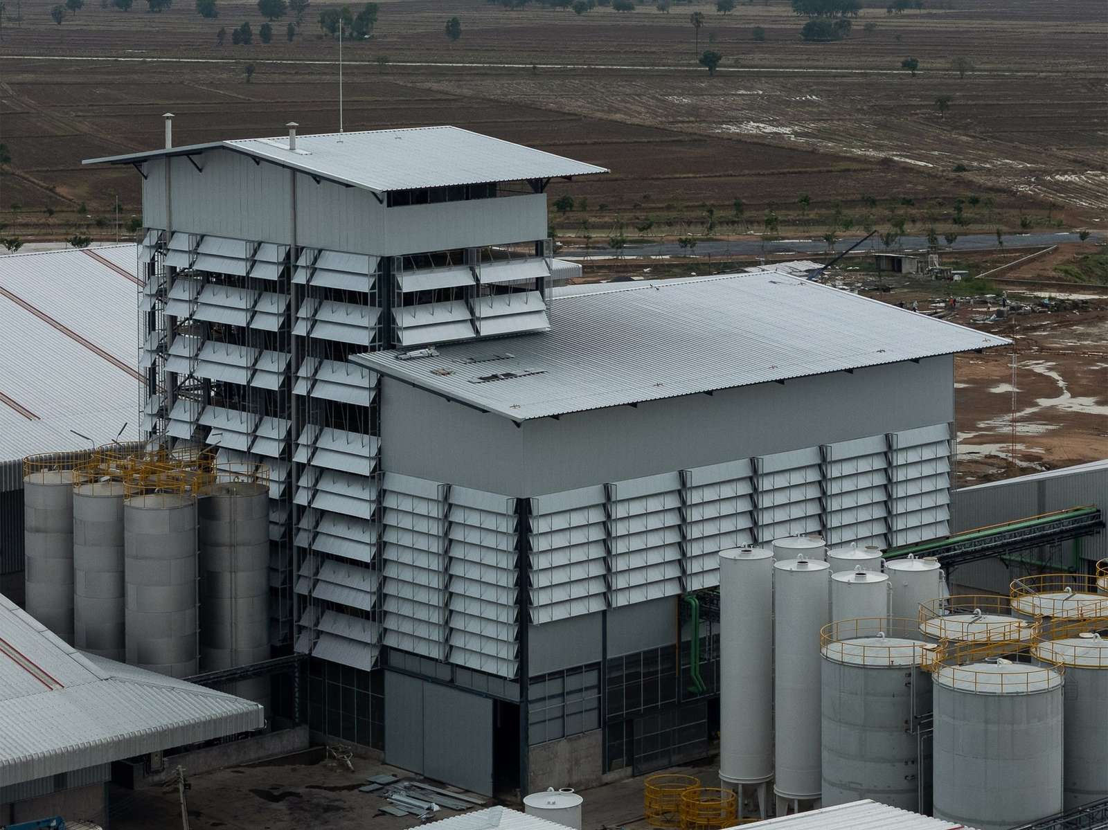 Aerial shot of an industrial rice bran oil refinery facility featuring corrugated metal facade louvers and storage silos set against open agricultural fields.