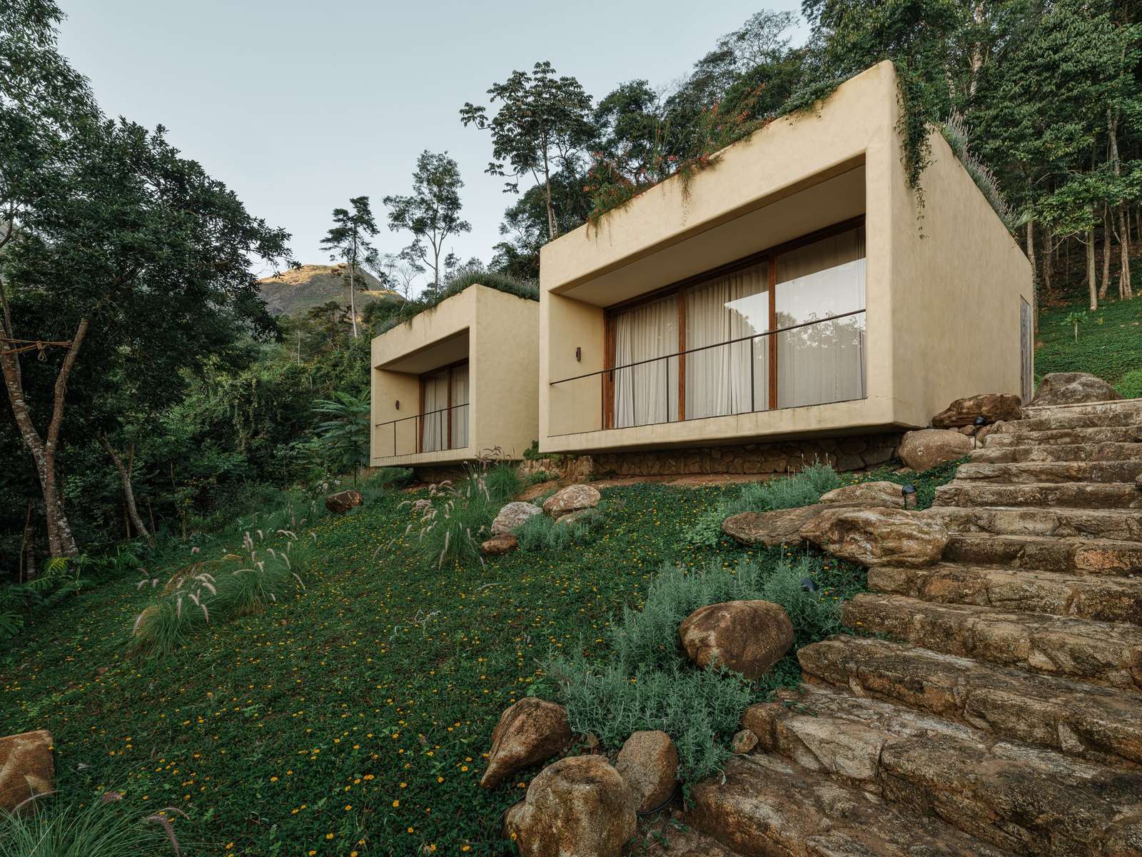 Exterior view of concrete suite volumes with green roofs nestled into a grassy mountain slope at Japú House by AIA Estúdio.