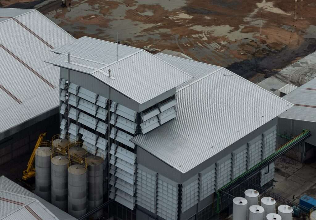 High-angle aerial view of the industrial plant showing sloped corrugated metal roofs, dynamic louvered facades, and vertical storage tanks.