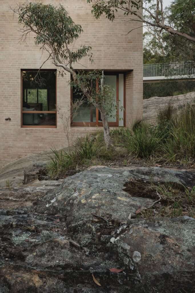A close-up view of EA House’s light brick facade featuring a large wooden-framed window and a pedestrian bridge on the right, with rugged, lichen-covered bedrock in the foreground.