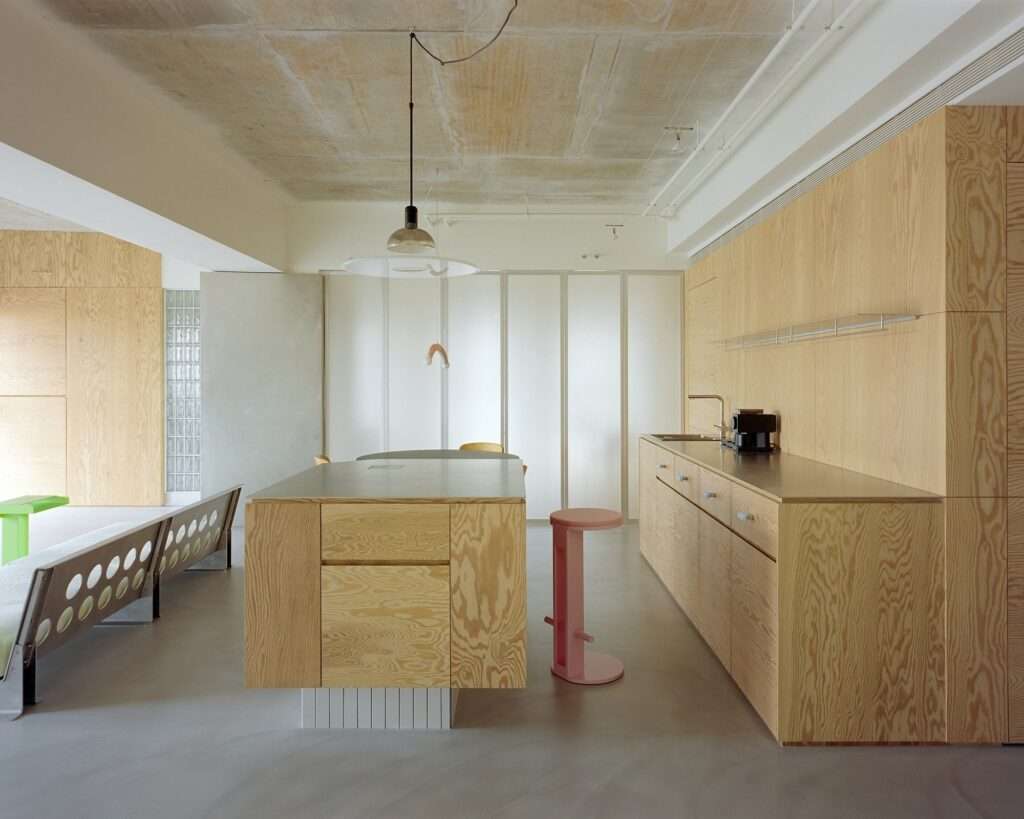 Open-plan kitchen in Raw and Warm Residence featuring pine plywood cabinetry, island counter, exposed raw concrete ceiling, smooth grey floor, and a pink bar stool accent.
