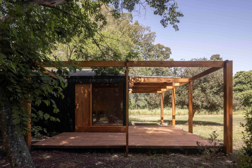 Exterior view of Etchegoyen House showing timber deck, wooden pergola frame, black wooden cladding, and lush green trees.