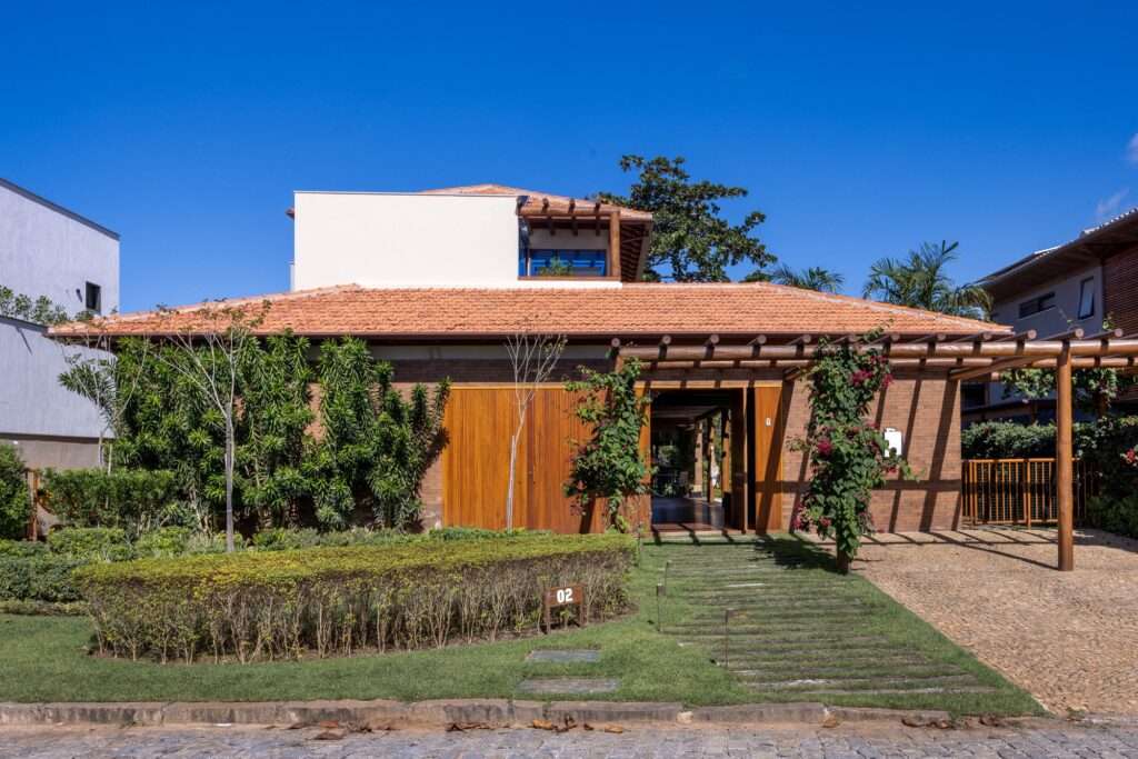 Front facade of MFA House featuring exposed brick walls, clay tile roof, eucalyptus timber pergola over driveway, and lush tropical landscaping.