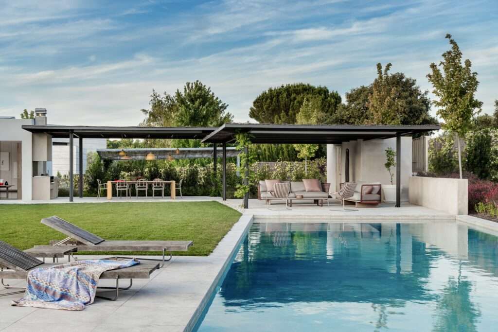 Wide shot of a luxury outdoor swimming pool terrace at Naseem House, featuring light gray paving, wooden sun loungers with a patterned blanket, a covered dark metal pergola sheltering a lounge area, and an outdoor dining space surrounded by greenery.