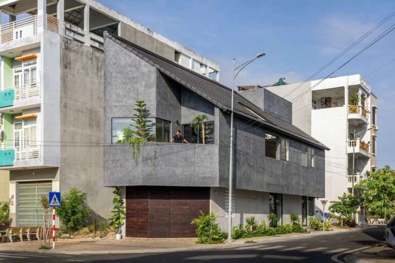 Exterior view of the Inner Courtyard House in Phan Rang on a sunny day, showcasing a dark exposed aggregate concrete facade, sloped tile roof, and wooden entrance gate on a corner plot.