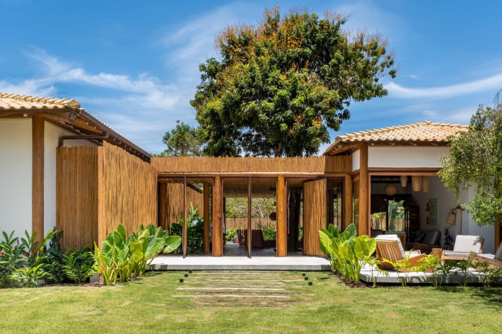 Main entrance of Ichiguin House in South Bahia featuring eucalyptus wood columns, biriba screens, and tropical vegetation under a sunny sky.