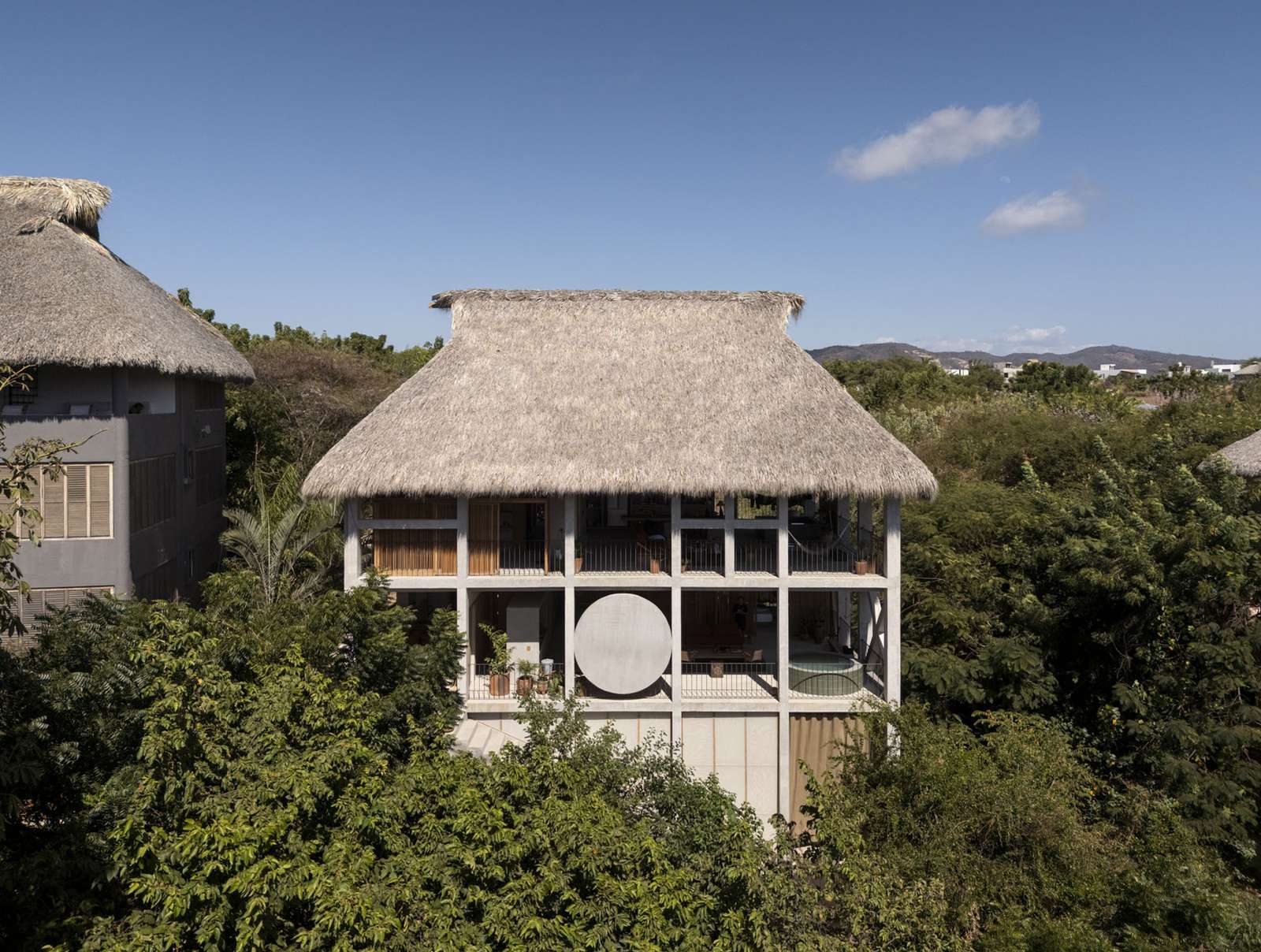 Front view of Casa Eva showing the exposed concrete structural grid, circular wall element, open terraces, and thatched palapa roof surrounded by tropical foliage.
