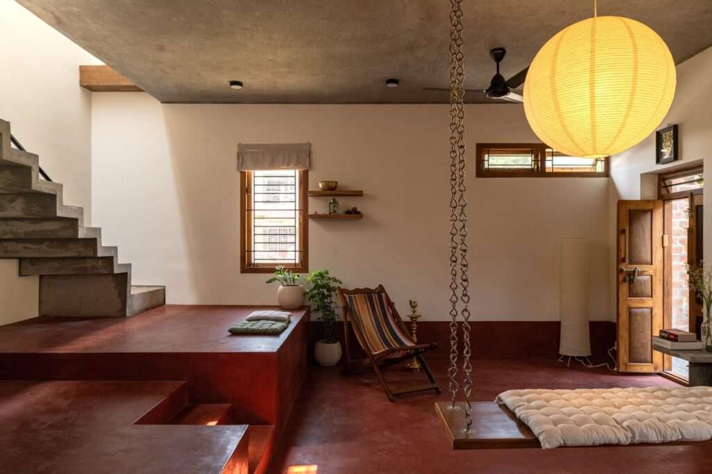 Ground floor interior of Thuraiyur Farmhouse, featuring red oxide flooring, a traditional wooden swing with cushions, a large spherical paper lantern, and an exposed concrete staircase, showcasing a flexible living area.