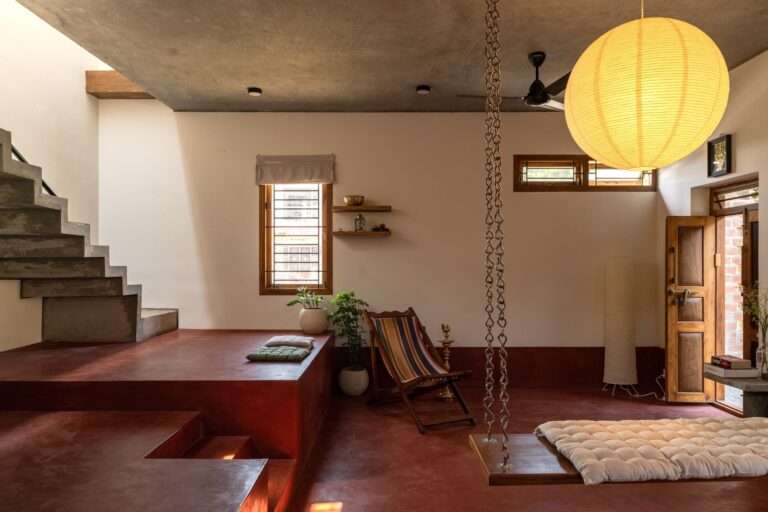 Ground floor interior of Thuraiyur Farmhouse, featuring red oxide flooring, a traditional wooden swing with cushions, a large spherical paper lantern, and an exposed concrete staircase, showcasing a flexible living area.