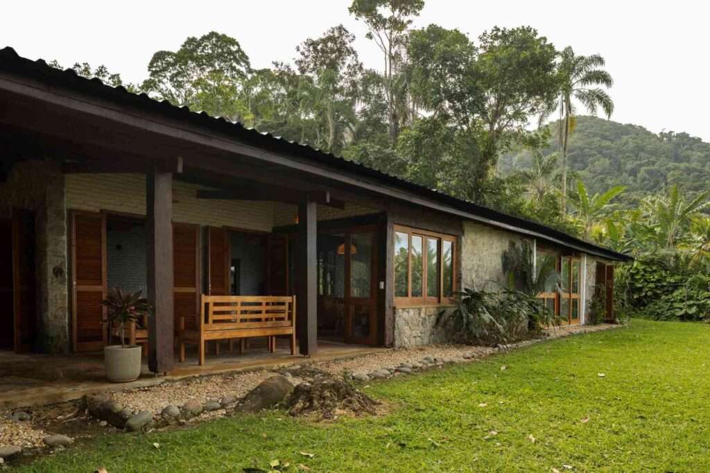 Exterior view of a coastal house in São Paulo showing white-painted brick, stone walls, wooden porch columns, and a lush green lawn surrounded by tropical forest.