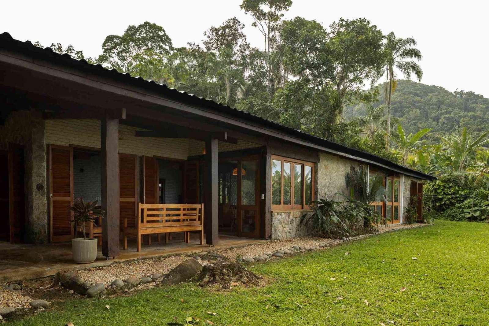 Exterior view of a coastal house in São Paulo showing white-painted brick, stone walls, wooden porch columns, and a lush green lawn surrounded by tropical forest.