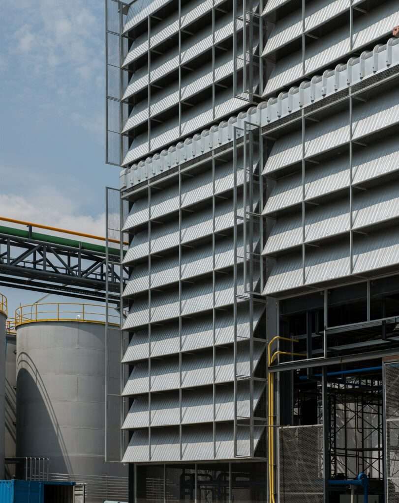 Close-up exterior view of steel-framed louvers and wire mesh screens mounted on an industrial factory elevation beside storage tanks.