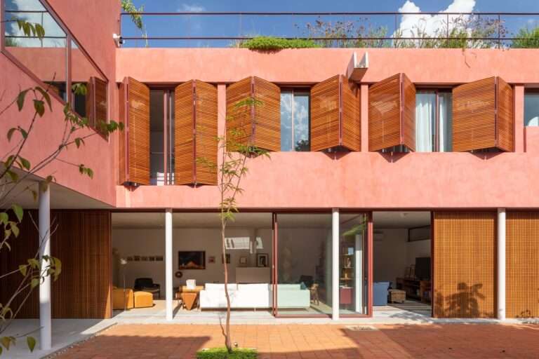 L-shaped house courtyard view featuring pink exterior walls, lower-level sliding wooden screens, upper-level folding louvers, and a brick-paved patio.