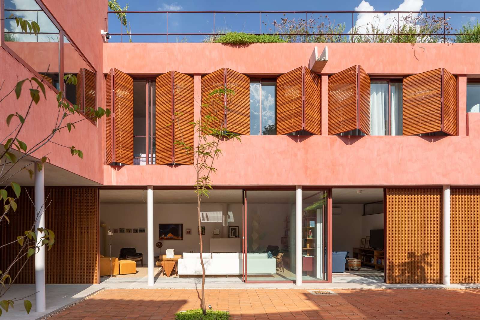 L-shaped house courtyard view featuring pink exterior walls, lower-level sliding wooden screens, upper-level folding louvers, and a brick-paved patio.