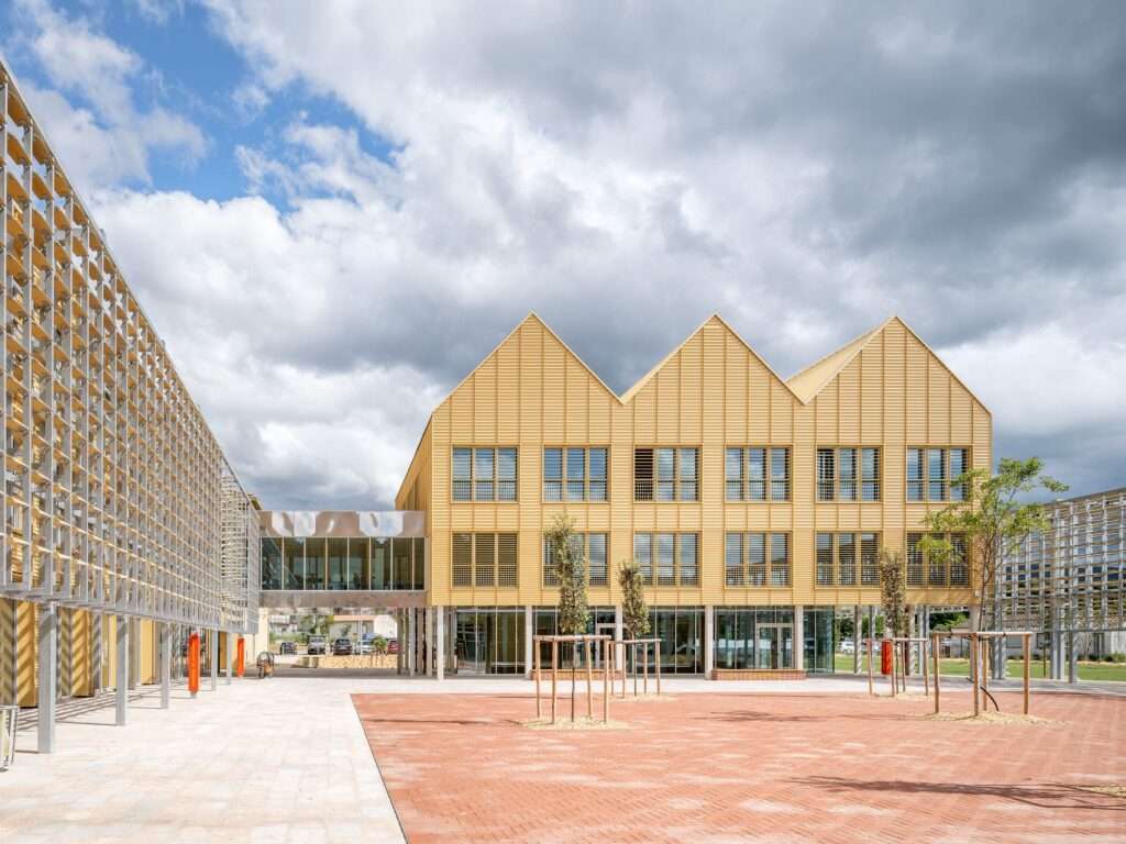 Exterior view of La Tuilerie cultural centre in Limoux designed by Ferrier Marchetti Studio showing yellow saw-tooth roof structures, brick paved public square, and terra-cotta sunshade screen.