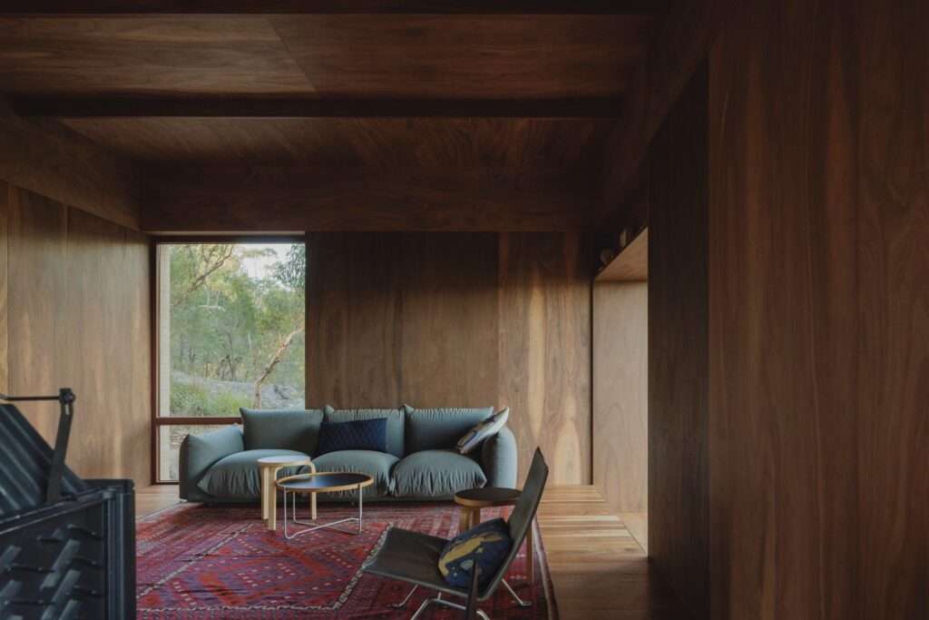 The interior of EA House’s living area, featuring dark timber plywood-paneled walls and ceiling, a blue sofa on a red patterned rug, a leather chair, and a view of the bushland through a single window.
