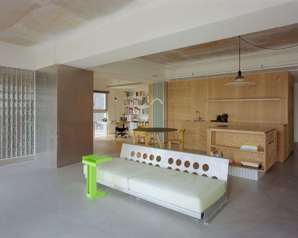 Modern living room in Raw and Warm Residence with perforated metal sofa, fluorescent green side table, pine plywood kitchen cabinetry, and glass block wall partition.
