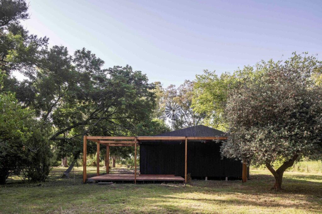 Etchegoyen House dark wooden volume nestled in a green clearing surrounded by trees and a traditional windmill in the background.
