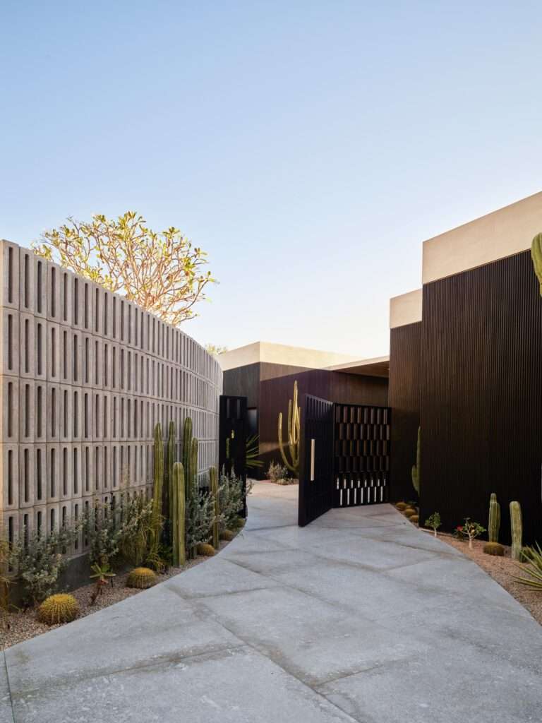 Concrete breeze block screen wall leading along a paved entrance path to dark textured concrete house walls and desert cacti.