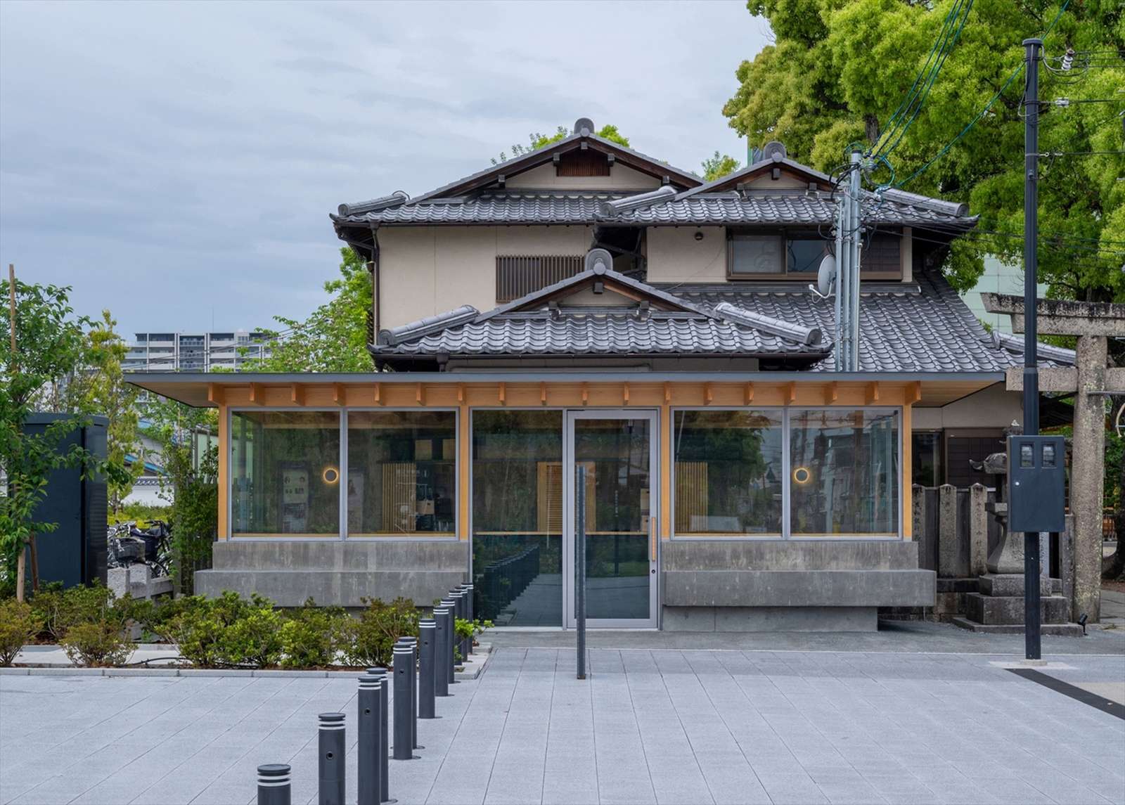 Front view of Cafe N+ featuring a modern glass facade, concrete plinth, and timber roof extension built under a traditional Japanese tiled roof building next to a stone shrine gate.