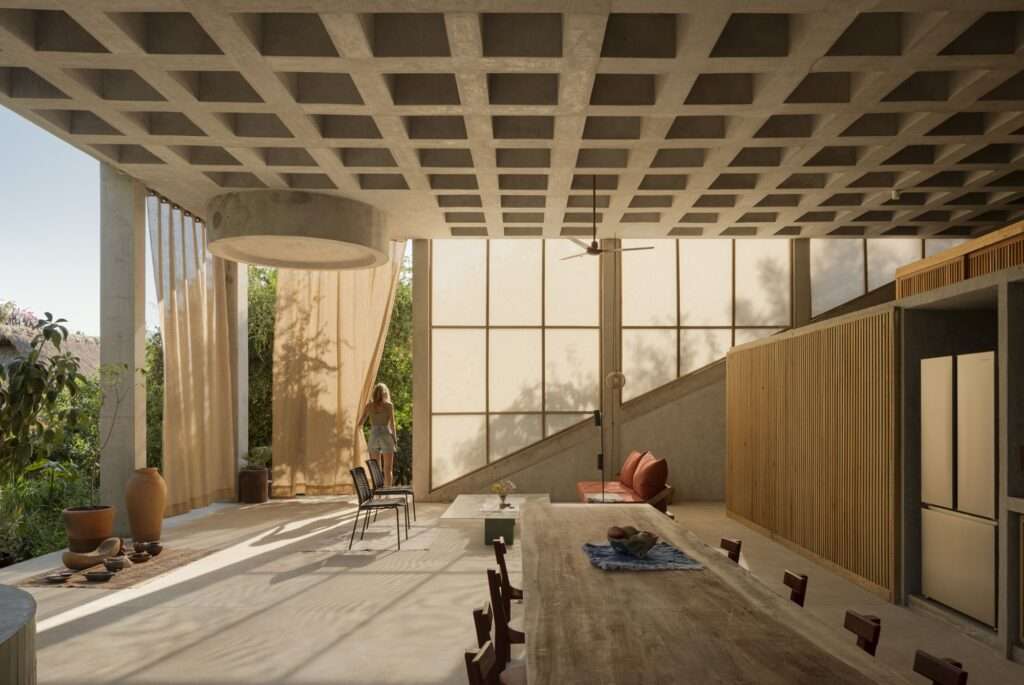 Open-plan living interior under a coffered concrete ceiling with a long wooden dining table, sheer curtains, and natural light filtering through tall glass panels.