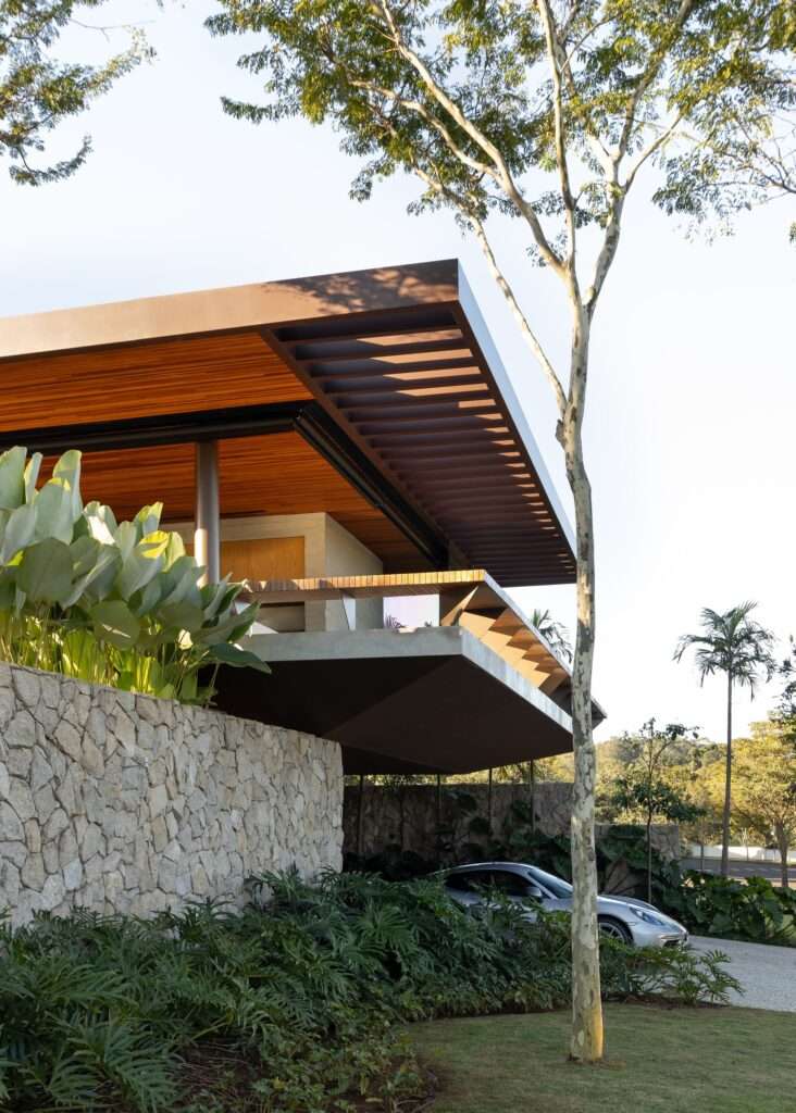 Vertical close-up architectural view of Casa da Mata showing a slatted roof overhang, raw stone wall, and native trees framing the building.