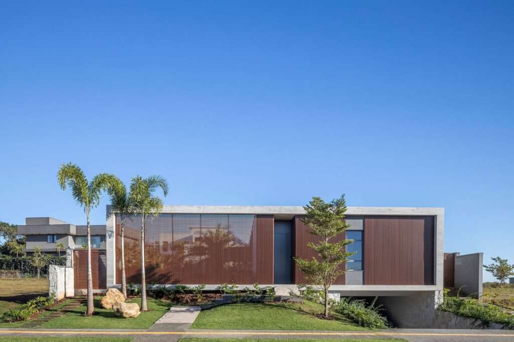 Modern main facade of Jabuticabeiras House featuring an exposed concrete frame, wood-look aluminum mashrabiya louvers, and manicured tropical landscaping.