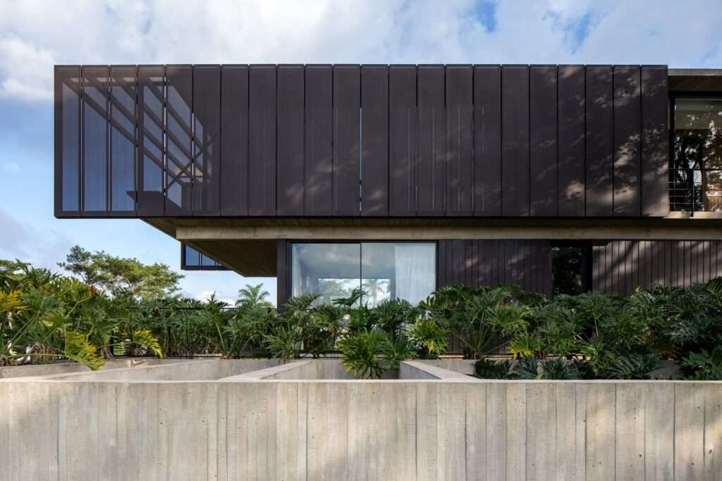 Cantilevered upper volume of Casa Galería clad in dark perforated metal panels extending over a concrete base surrounded by lush green foliage.