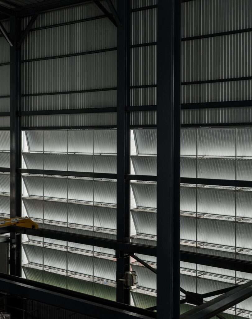Interior perspective looking out toward permeable facade panels, showing diffused light and exposed structural steel frames in a production hall.