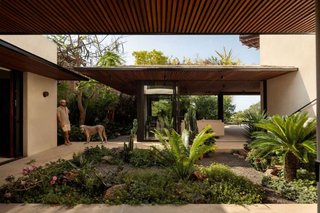 Central courtyard of a modern home featuring a desert-style garden, covered walkways with wooden slatted ceilings, and a view extending towards a pool.