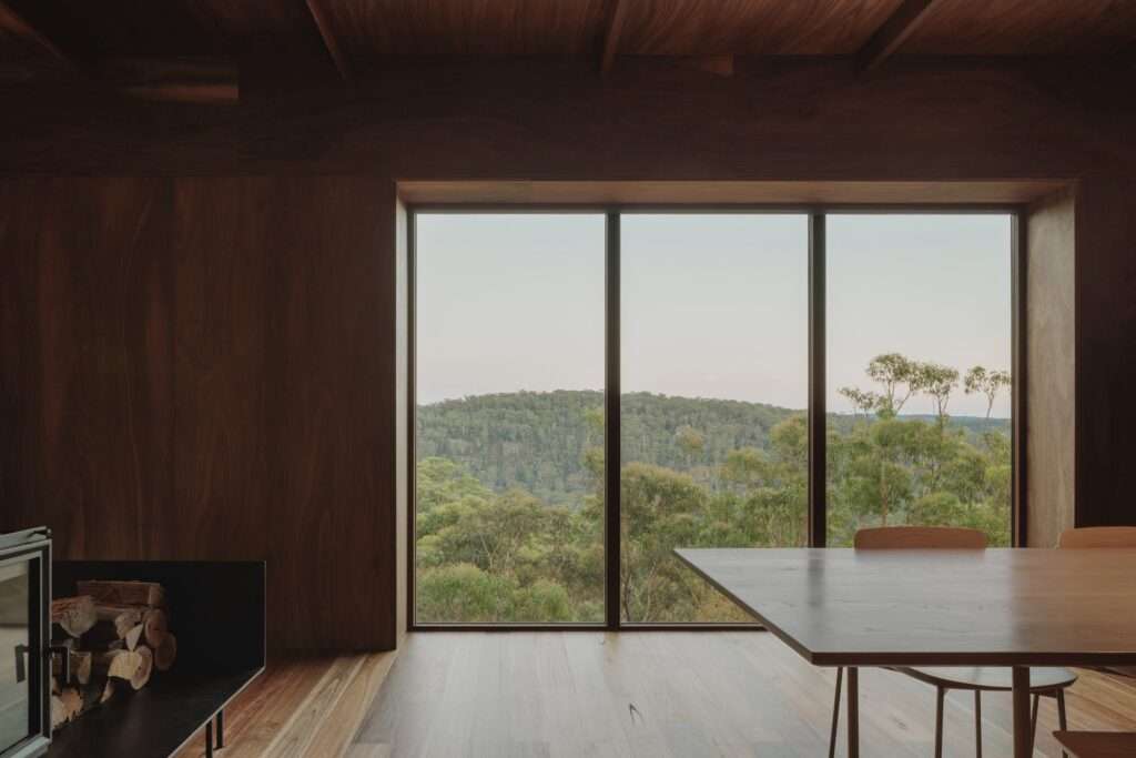 The lower-level dining area of EA House, showing a timber table and chairs in the foreground, polished wooden floors, dark wood-paneled walls, and a large three-panel window offering an expansive view of a dense, green valley.