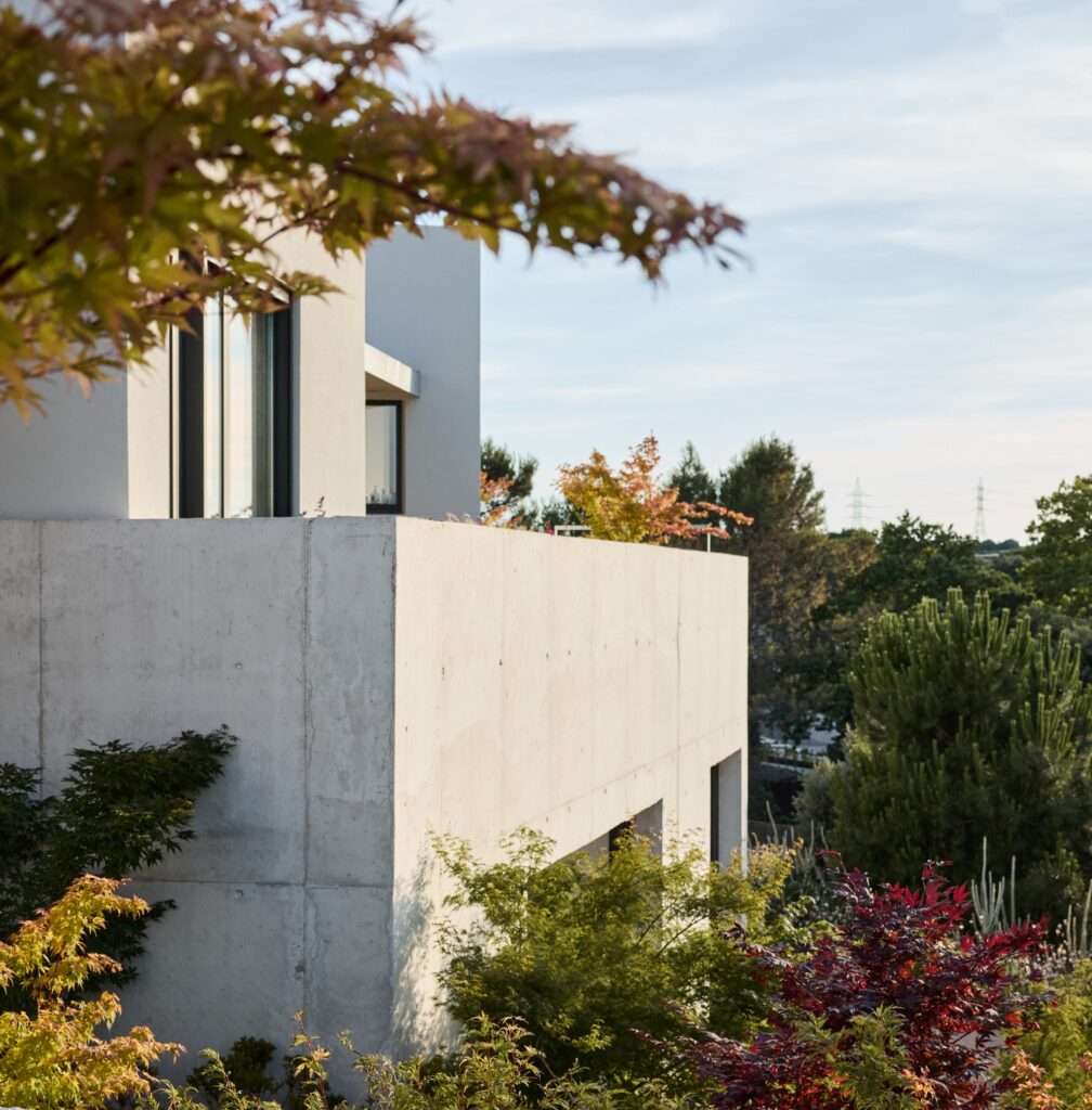 External architectural shot showing the raw concrete base of Naseem House embedded in a hill with red and green foliage, topped by a white upper floor volume and dark-framed windows under a clear sky.