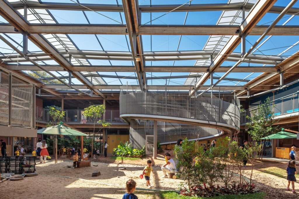 Glulam timber frame structure over a central courtyard with a central helical ramp, sand pit play area, and natural translucent roof panels.
