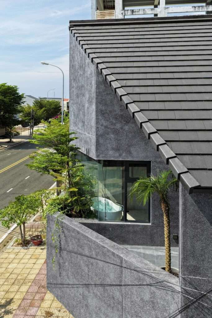 High-angle view looking down at the dark tiled pitched roof, textured concrete walls, and an upper-floor outdoor terrace with tropical plants and a bathtub visible through glass.